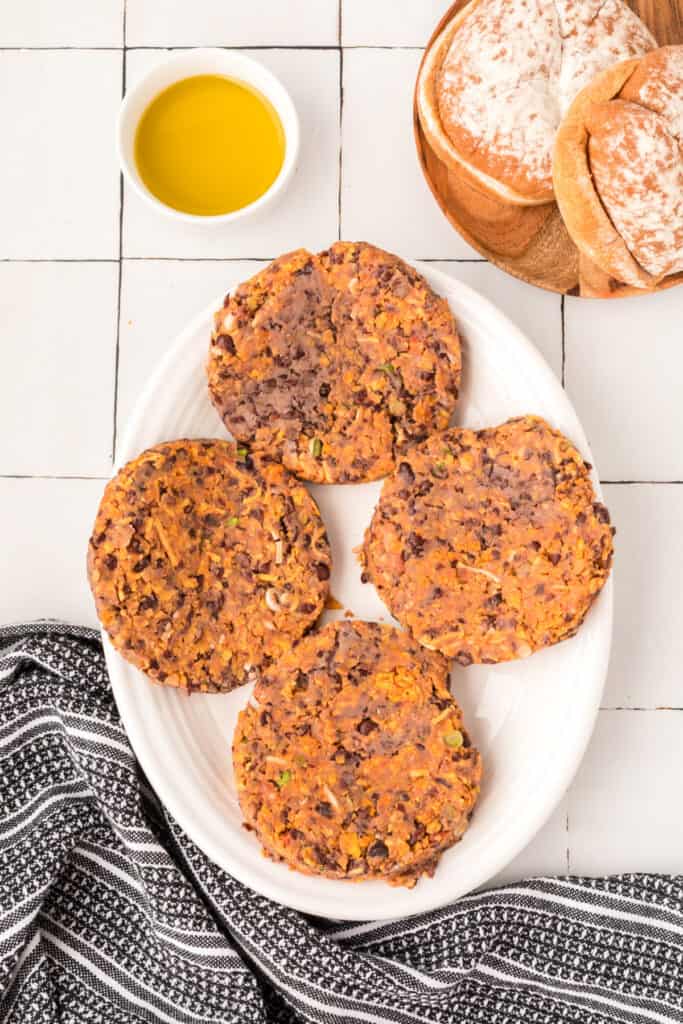 four uncooked Black Bean Nacho Burger patties shaped on a white plate next to buns and oil