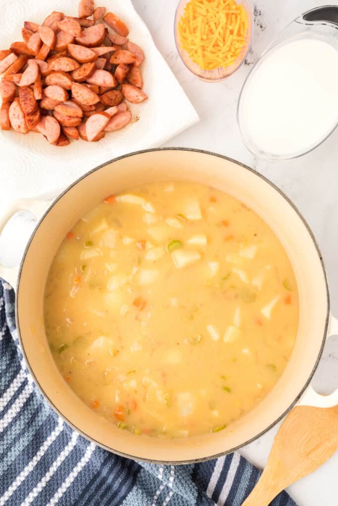 Soup simmering in a large pot with potatoes and vegetables, with browned sausage, shredded cheese, and milk set aside.