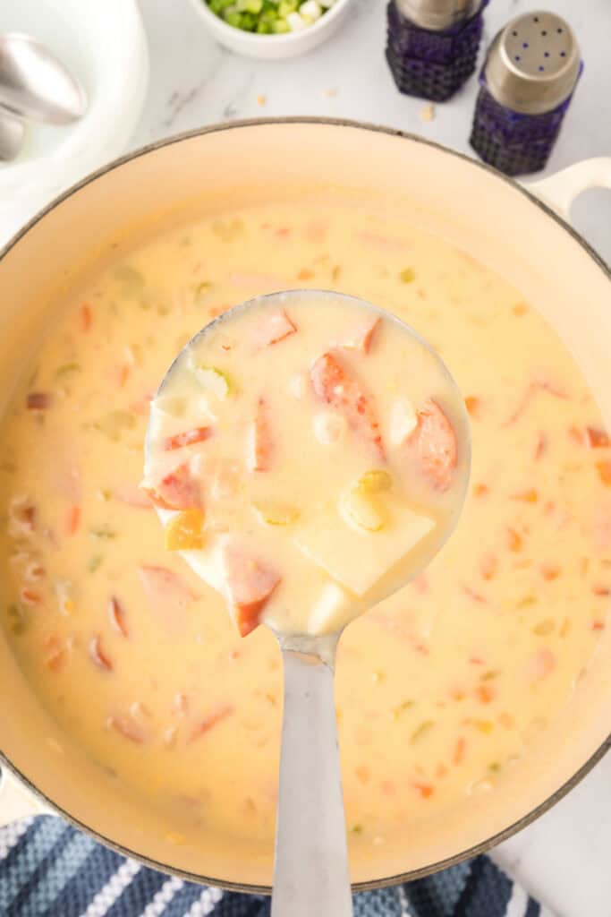 Ladle lifting a hearty serving of cheesy kielbasa potato soup filled with sausage, potatoes, and vegetables.