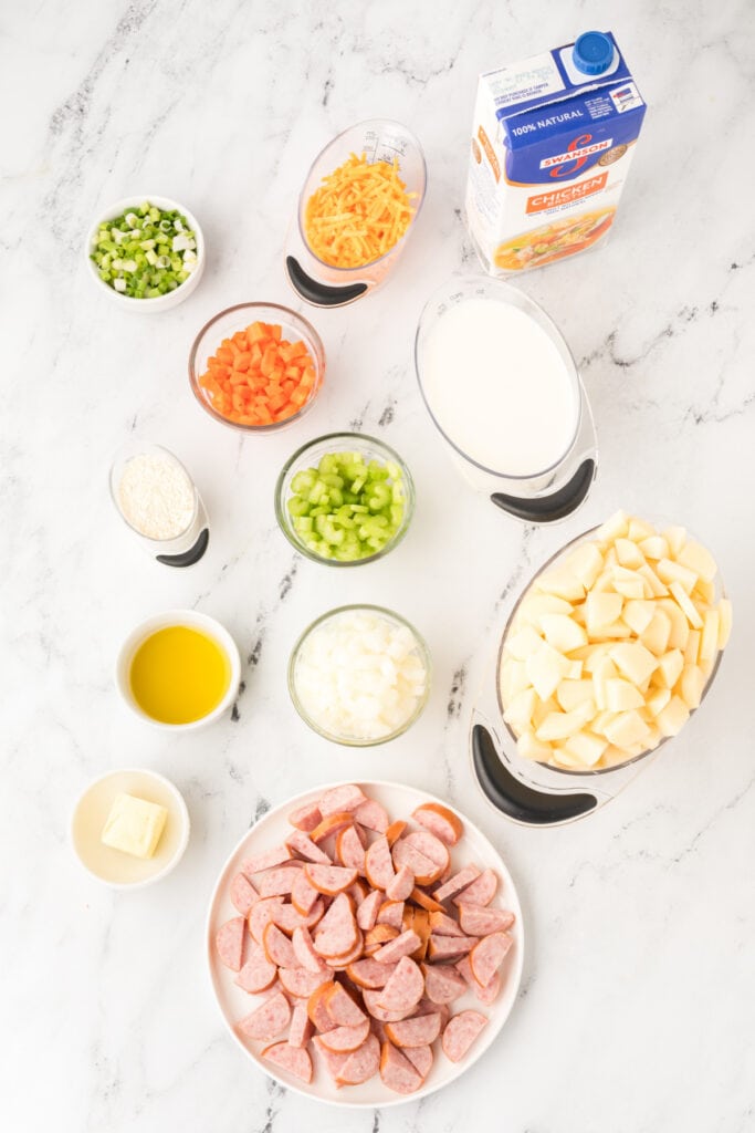Overhead view of prepped ingredients for cheesy kielbasa potato soup including chopped veggies, diced potatoes, sliced sausage, broth, cheese, milk, butter, and flour on a marble surface.