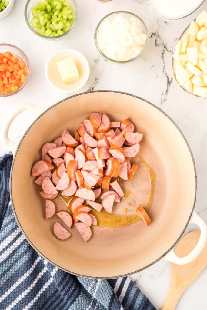 Sliced kielbasa sausage added to a large soup pot with oil, surrounded by bowls of chopped vegetables and potatoes.