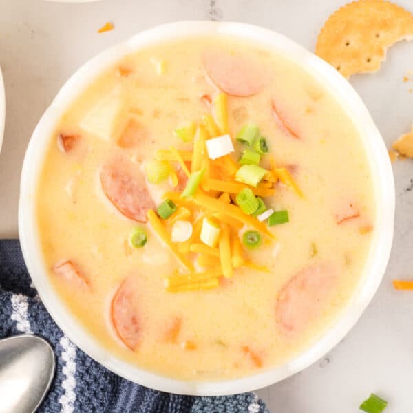 Bowl of cheesy kielbasa potato soup topped with shredded cheddar and green onions, served with crackers on the side.
