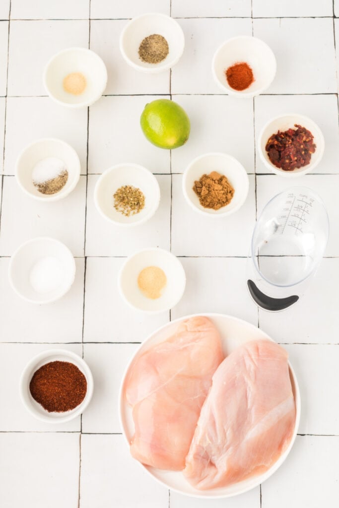 Ingredients for chipotle lime chicken tacos arranged on a white tiled surface with chicken breasts, spices, lime, chipotle peppers, and water