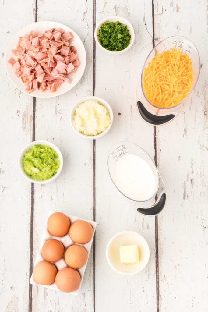 Overhead view of diced ham, shredded cheddar, chopped parsley, eggs, milk, butter, and grated onion arranged on a white rustic surface.