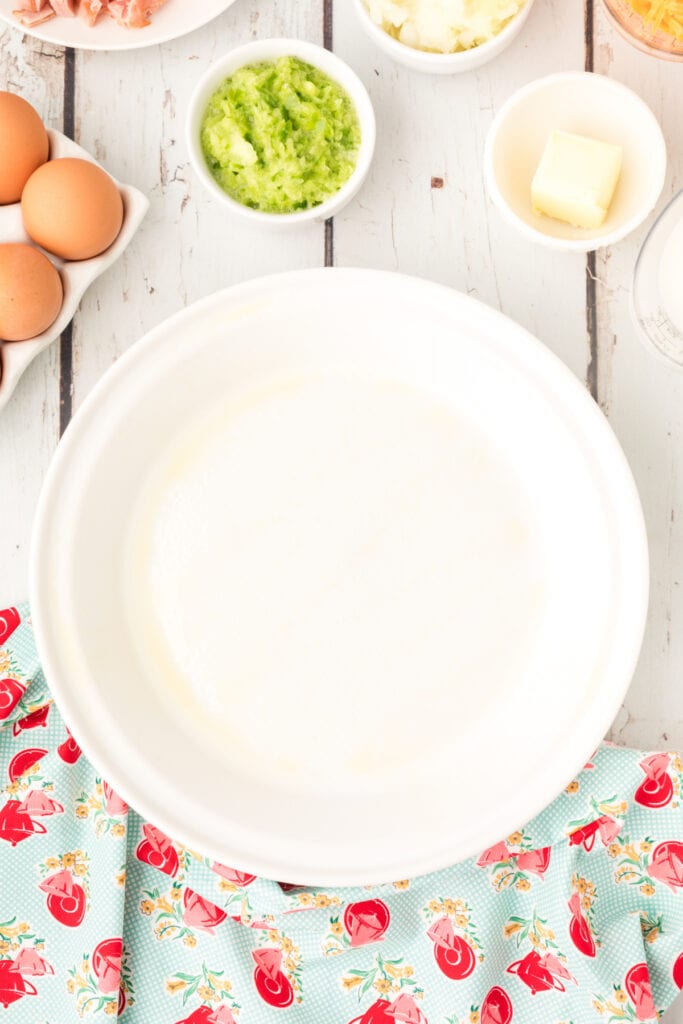 Greased pie plate and small prep bowls arranged on a white wooden surface with eggs, grated vegetables, butter, and cheese nearby.