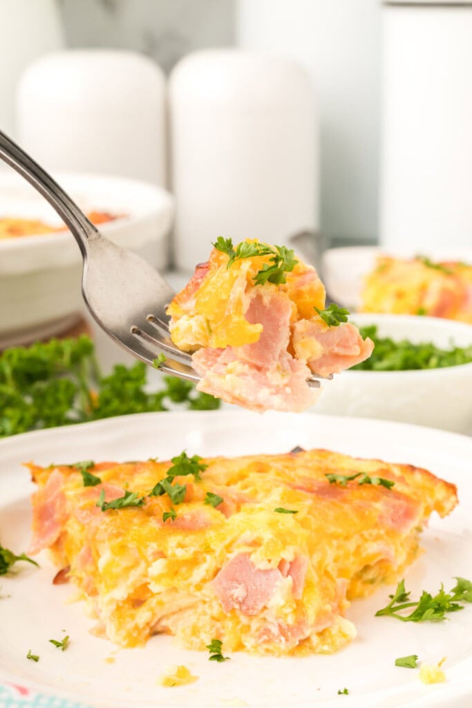 Fork lifting a bite of Crustless Ham Bake above a plated slice with parsley garnish.