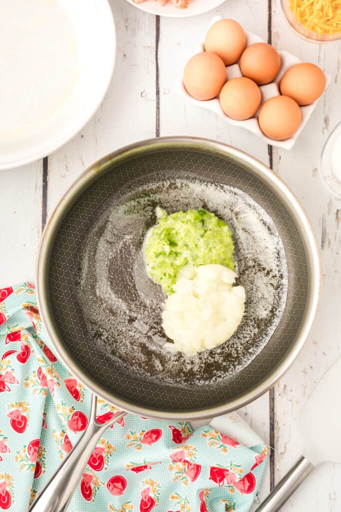 Skillet with melted butter holding grated onion and celery mixture on a rustic white background.