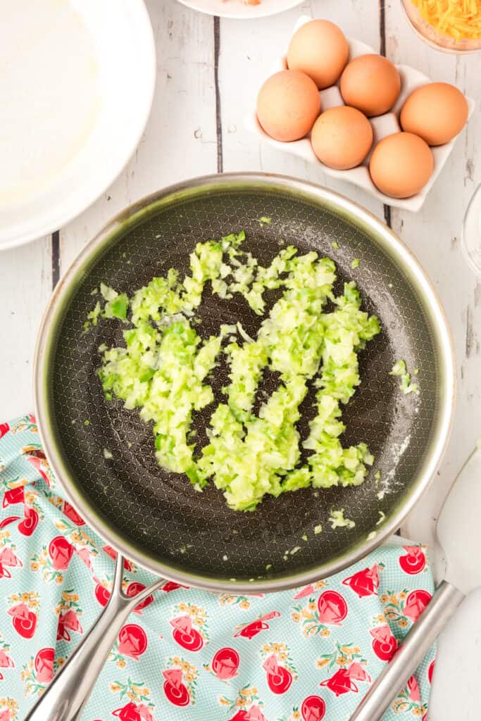 Sautéed onion and celery mixture cooking in a skillet on a white wooden surface.
