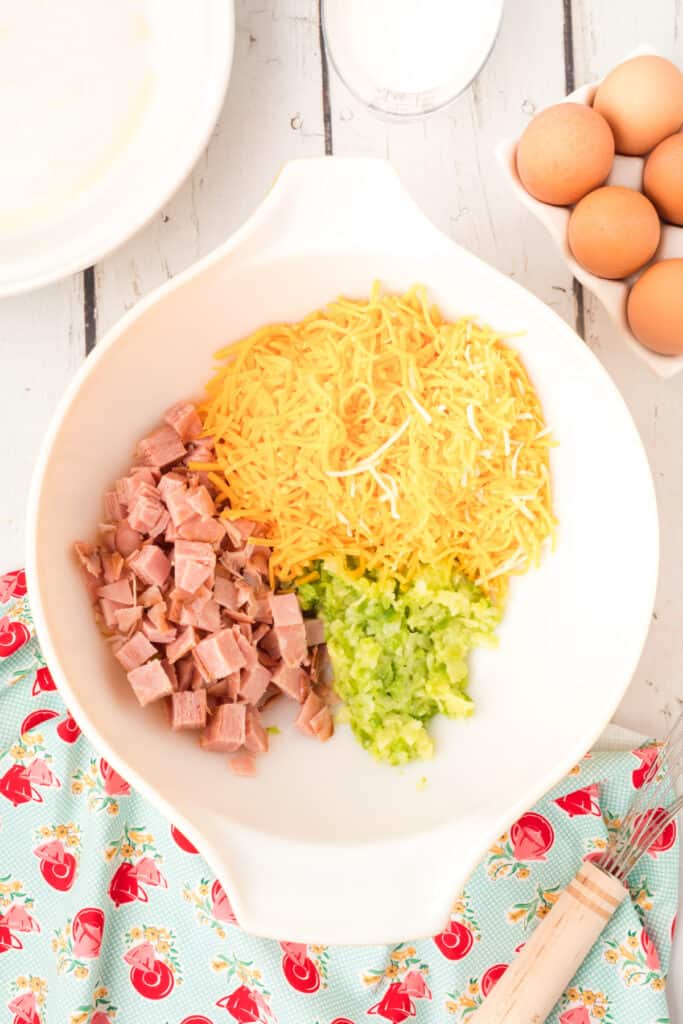 Large white mixing bowl filled with shredded cheddar, diced ham, and sautéed vegetables before mixing.