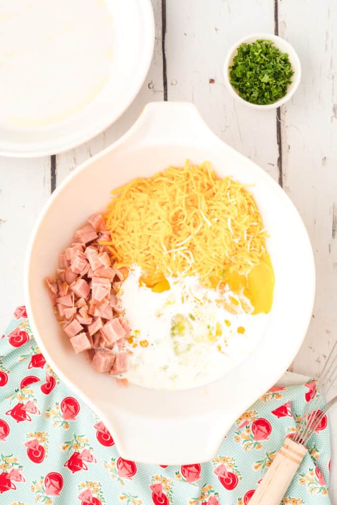 Mixing bowl holding shredded cheddar, diced ham, sautéed vegetables, eggs, and milk ready to be combined.