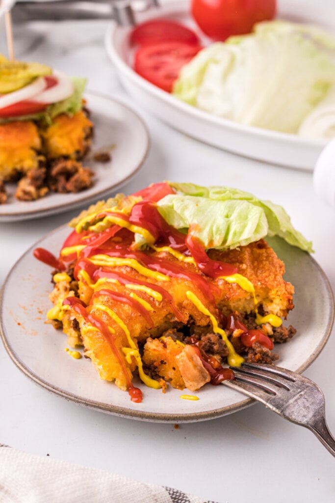 cheeseburger casserole topped with ketchup and mustard with lettuce tomato and pickles on the side