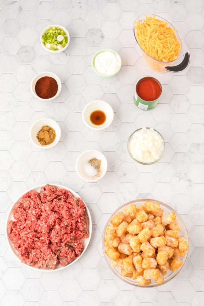 Overhead view of ingredients for Chili Cheese Tater Tot Casserole including ground beef, frozen tater tots, shredded cheddar cheese, tomato sauce, seasonings, onion, sour cream, and green onions on a white countertop.
