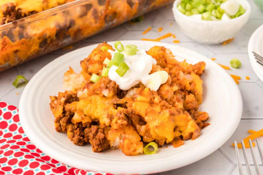 Wide view of a plated serving of Chili Cheese Tater Tot Casserole topped with sour cream and green onions beside the baking dish.