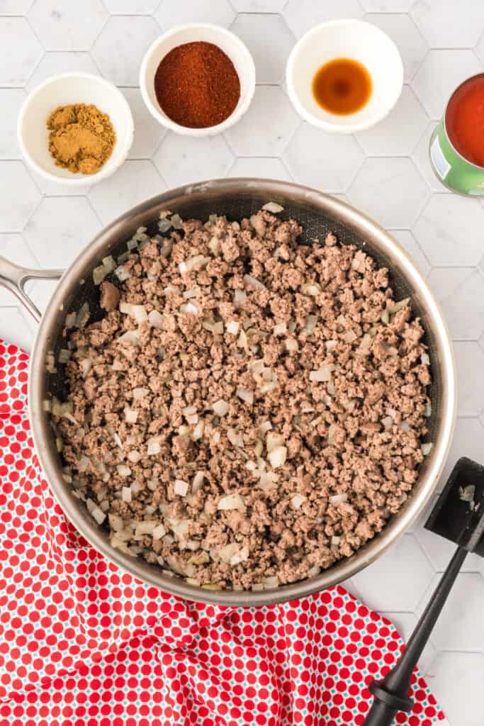 Cooked ground beef and onions in a skillet with bowls of chili powder, cumin, Worcestershire sauce, and tomato sauce nearby.