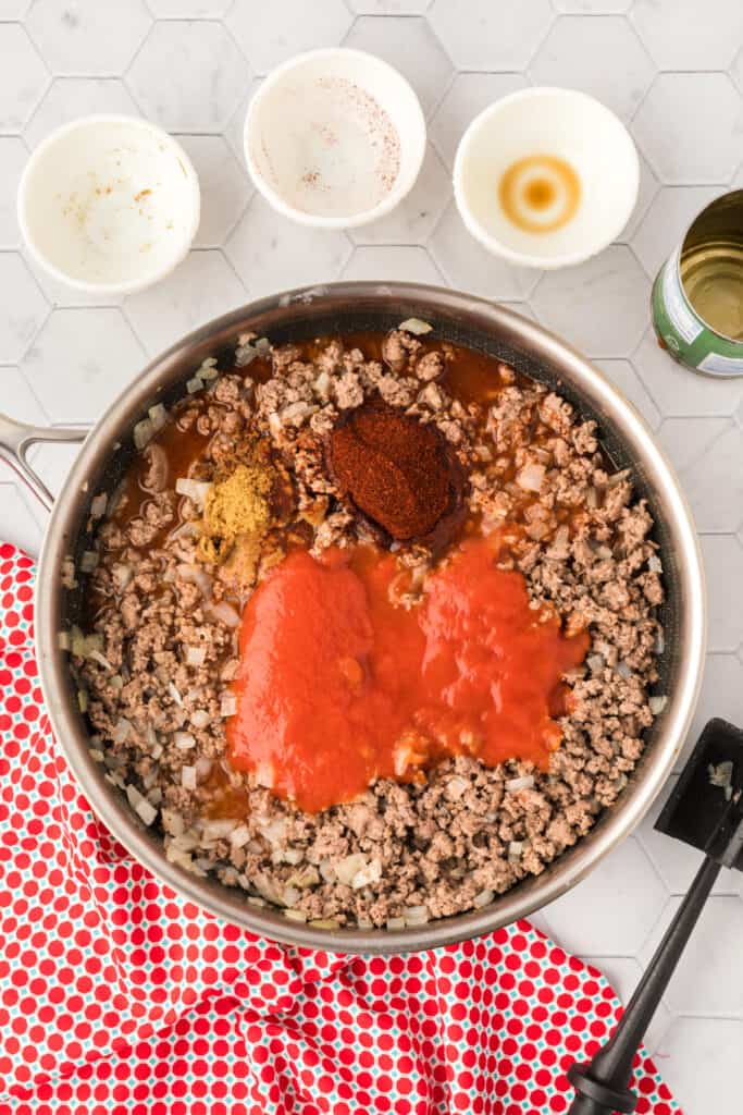 Cooked ground beef and onions in a skillet topped with tomato sauce, chili powder, cumin, Worcestershire sauce, and water before stirring.