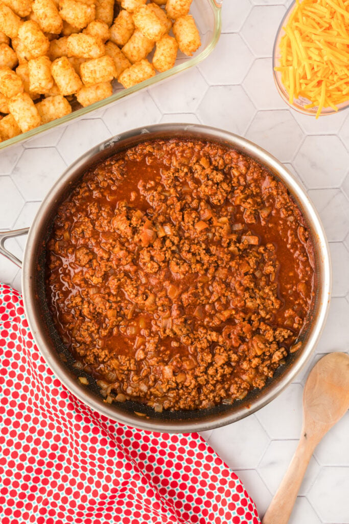 Homemade chili simmering in a skillet next to baked tater tots and shredded cheddar cheese.