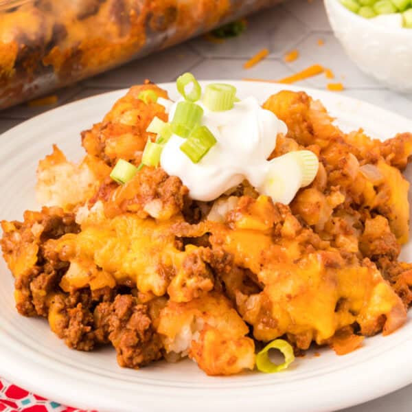 Square-cropped image of Chili Cheese Tater Tot Casserole topped with sour cream and sliced green onions on a white plate.