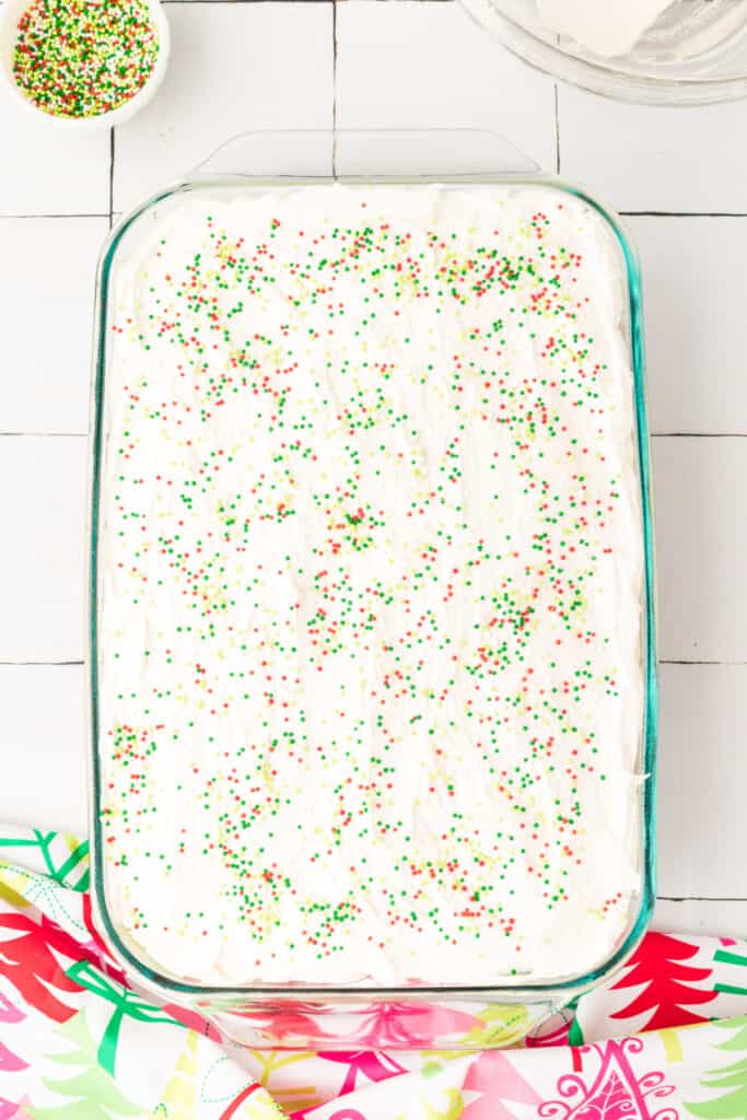 Red velvet poke cake in a glass baking dish topped with white cream cheese layer and scattered red, green, and white non-pareils.