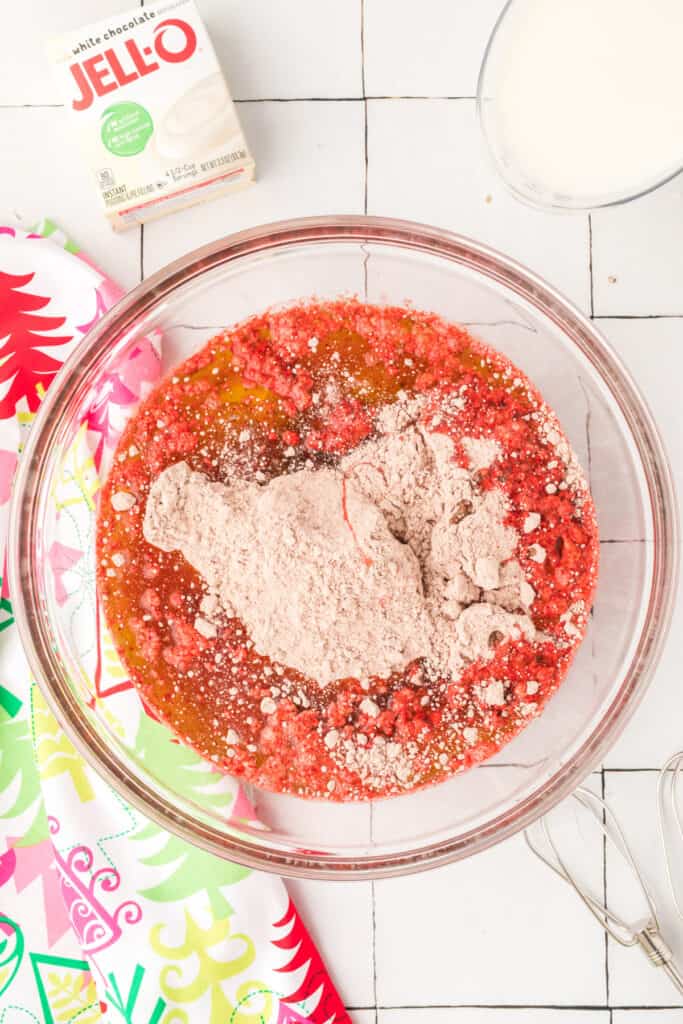 Overhead view of a glass mixing bowl with red velvet cake mix, eggs, oil, and dry ingredients before mixing. A box of white chocolate pudding and a bowl of milk sit nearby.