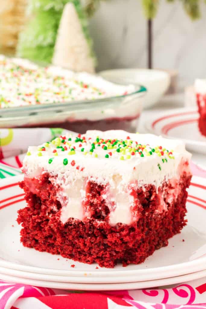Close-up of a slice of red velvet poke cake on a white plate with red stripes, showing pudding-filled holes and cream cheese topping with sprinkles.