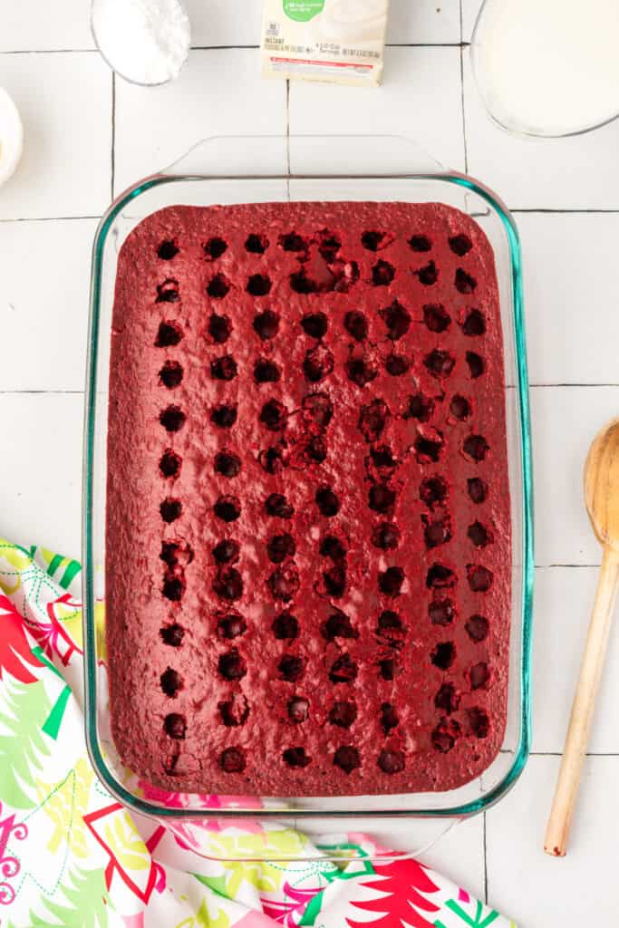 Baked red velvet cake in a glass baking dish with holes poked across the surface using the handle of a wooden spoon.