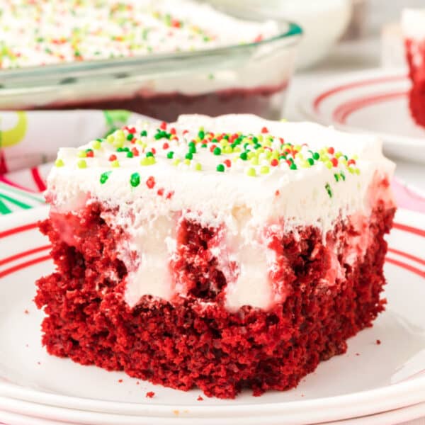 Square image of a slice of red velvet poke cake on a white plate with red stripes, topped with cream cheese layer and holiday sprinkles.