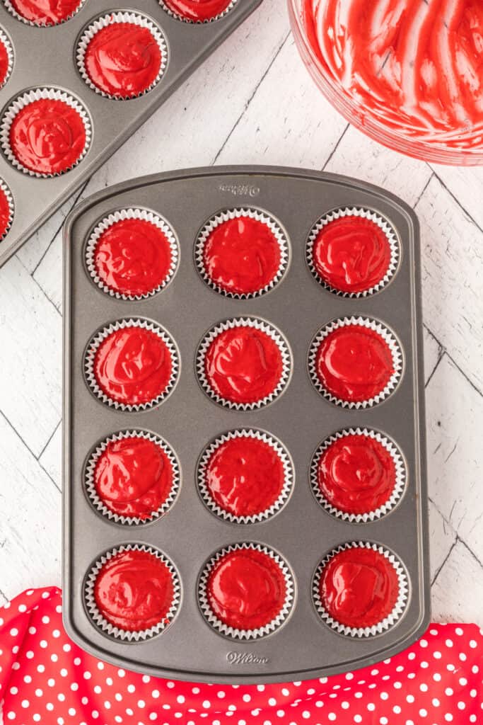 Muffin pan filled with white liners and bright red cupcake batter portioned into each cup, with extra batter in a bowl nearby.