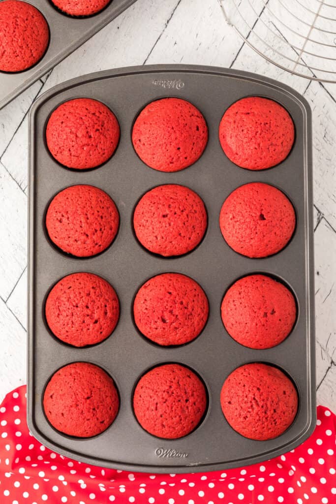 Baked red velvet cupcakes in a metal muffin pan with domed tops on a white wooden surface.