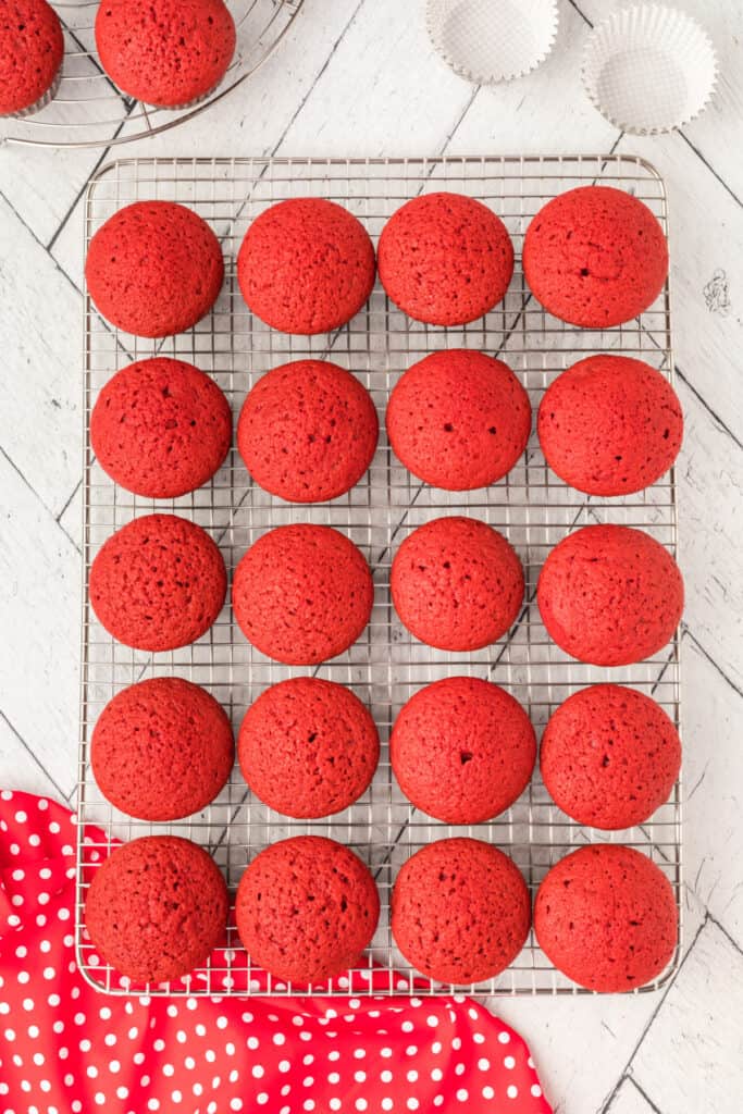 Twenty red velvet cupcakes cooling on a metal wire rack on a white wooden surface with a red polka dot cloth partially visible.