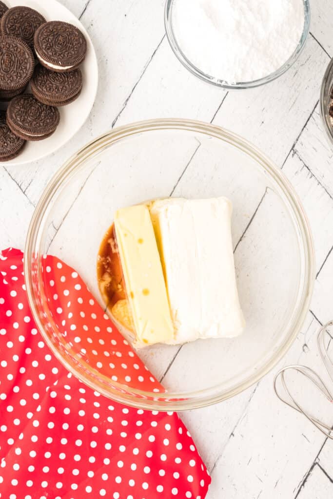 Glass mixing bowl with softened cream cheese and butter inside, with powdered sugar in a bowl and whole Oreos on a plate nearby.