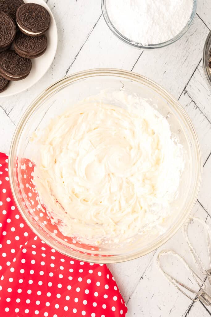 Glass mixing bowl with cream cheese and butter beaten smooth, with mixer beaters resting on the table beside it.