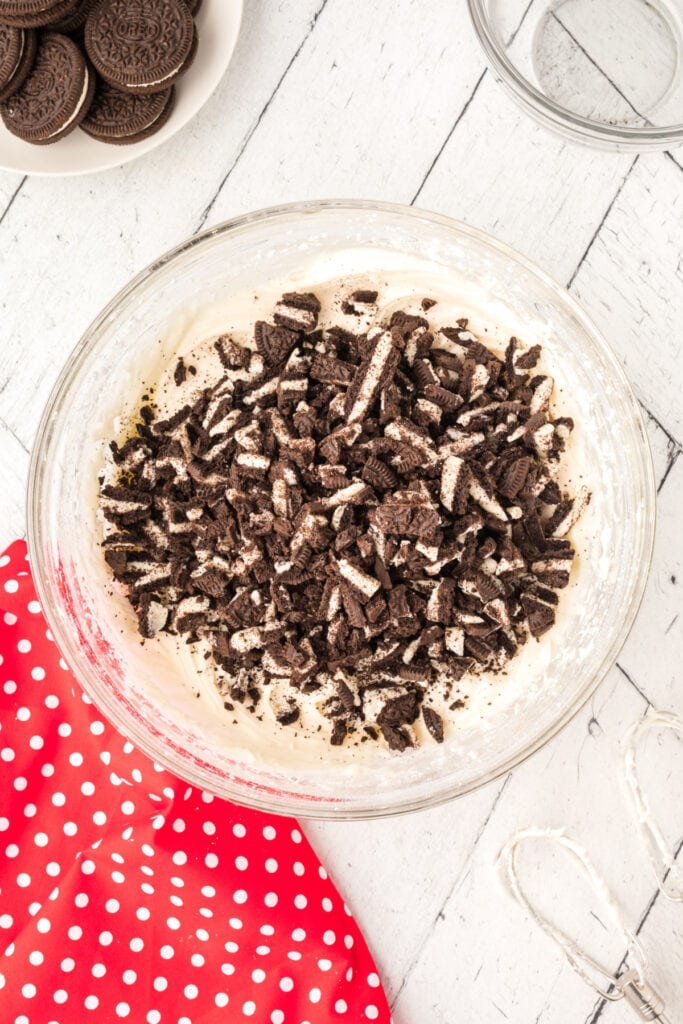 Glass mixing bowl filled with chopped Oreo pieces added on top of cream cheese frosting.