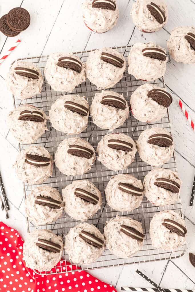 Red velvet cupcakes topped with Oreo cream cheese frosting arranged on a wire rack, each garnished with a halved Oreo cookie.