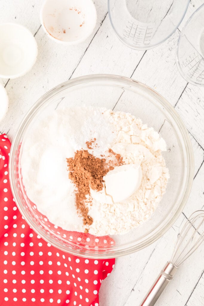 Glass mixing bowl with flour, sugar, cocoa powder, baking soda, and salt on a white wooden surface with measuring cups and a whisk nearby.