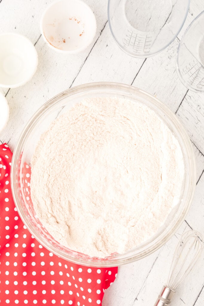 Glass mixing bowl with dry ingredients fully combined into a pale mixture on a white wooden surface with a whisk beside it.