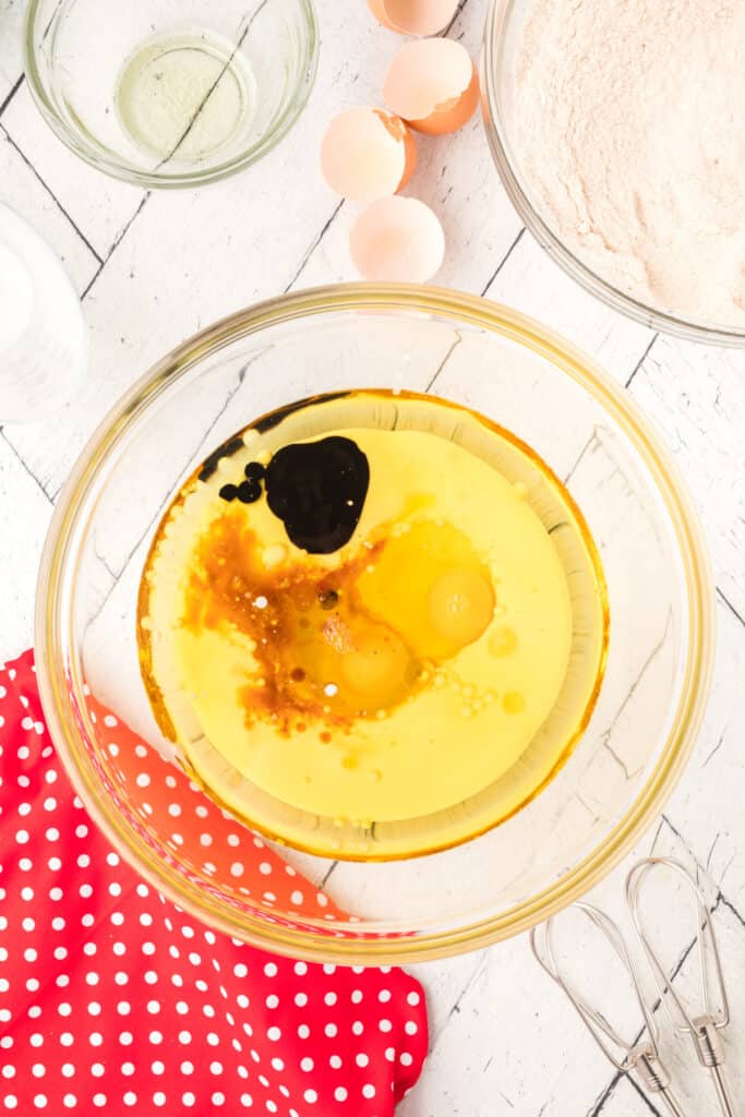 Glass bowl with oil, buttermilk, eggs, vanilla, vinegar, and red velvet emulsion before mixing on a white wooden surface.