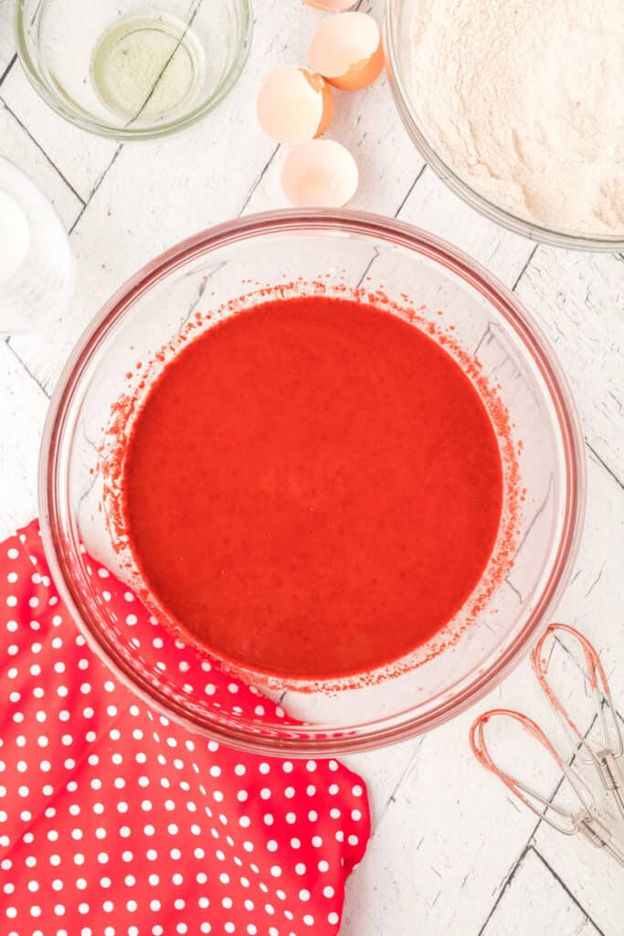 Glass bowl with bright red liquid batter mixture after wet ingredients have been combined on a white wooden surface.