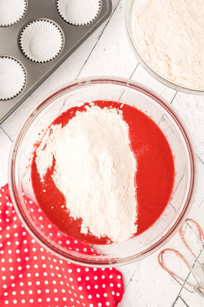 Glass bowl with dry ingredients added on top of red batter before mixing, with muffin pan and whisk nearby.