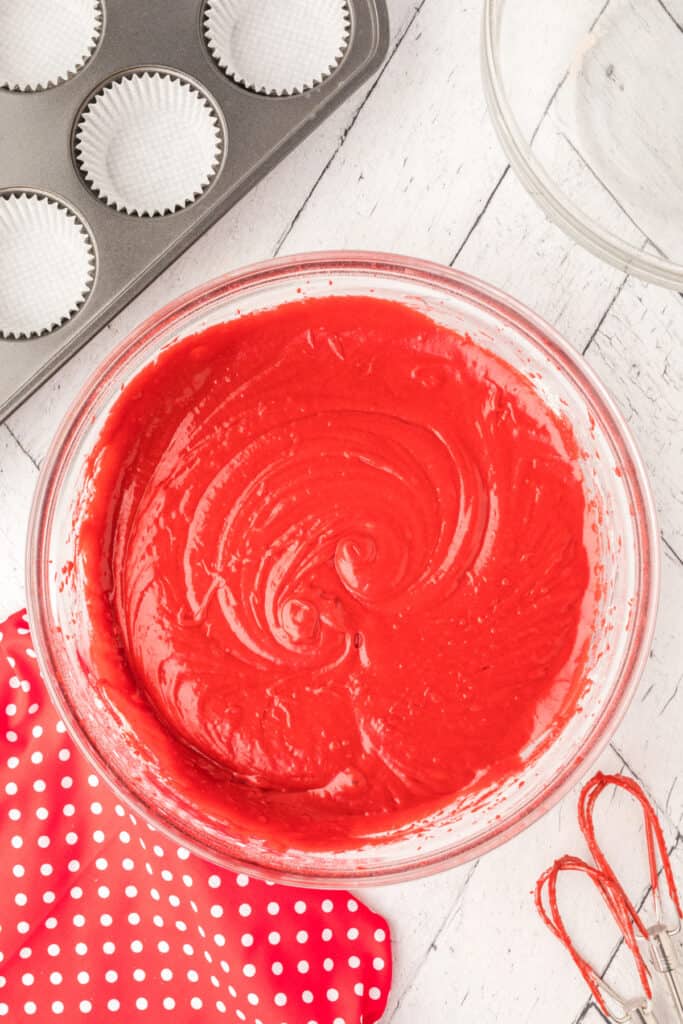 Glass bowl with smooth, fully mixed bright red cupcake batter on a white wooden surface.