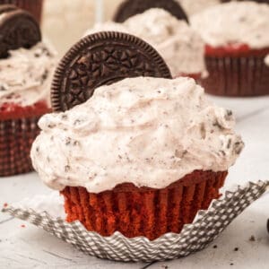 Close up of a red velvet cupcake with Oreo cream cheese frosting and a halved Oreo on top on a white surface.