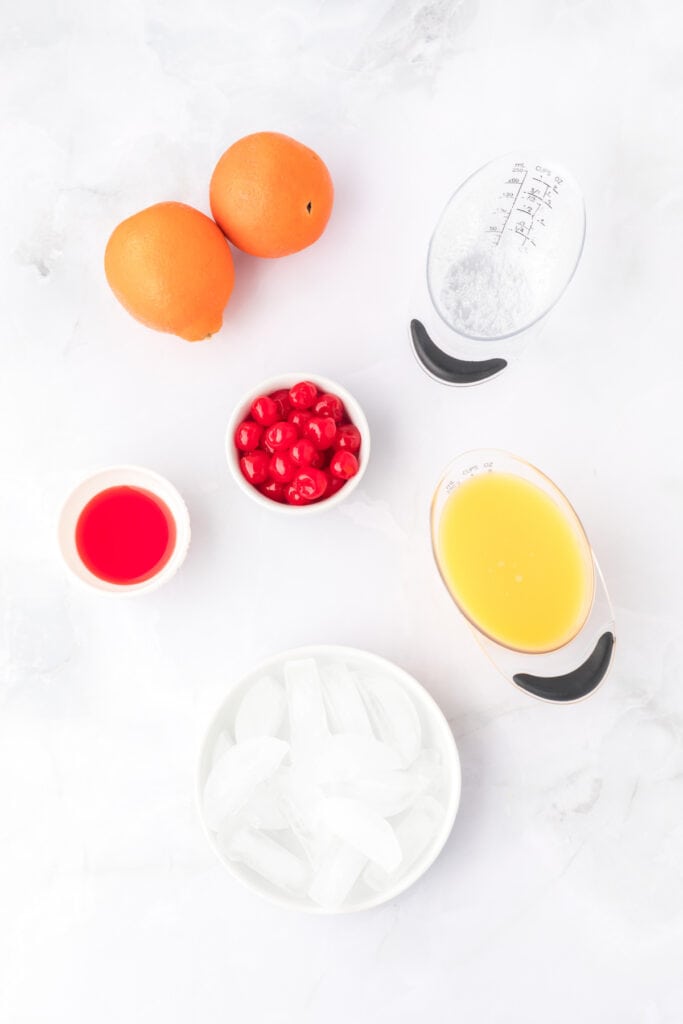 whole oranges, a measuring cup of orange juice, a measuring cup of clear soda, a small bowl of maraschino cherries, a small dish of grenadine, and a bowl of ice on a white surface