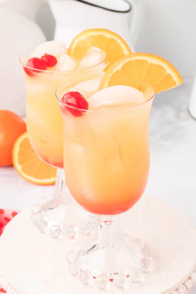 two glasses of layered orange and red drink with ice, garnished with orange slices and maraschino cherries