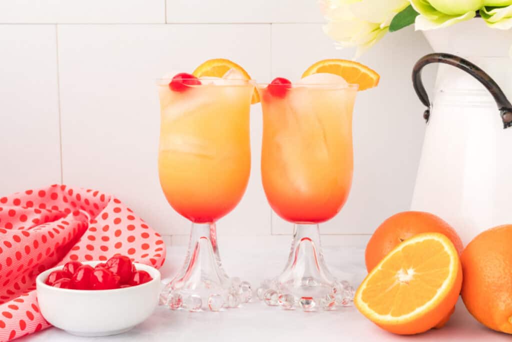 two glasses of layered orange and red drink with ice, garnished with orange slices and maraschino cherries