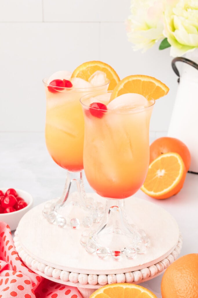 two Arizona Sunset drinks on a white stand with oranges and cherries nearby
