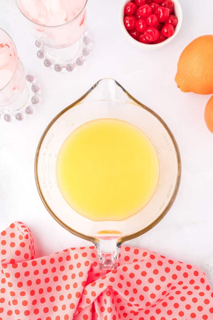 glass measuring cup filled with orange juice with glasses of ice and oranges nearby