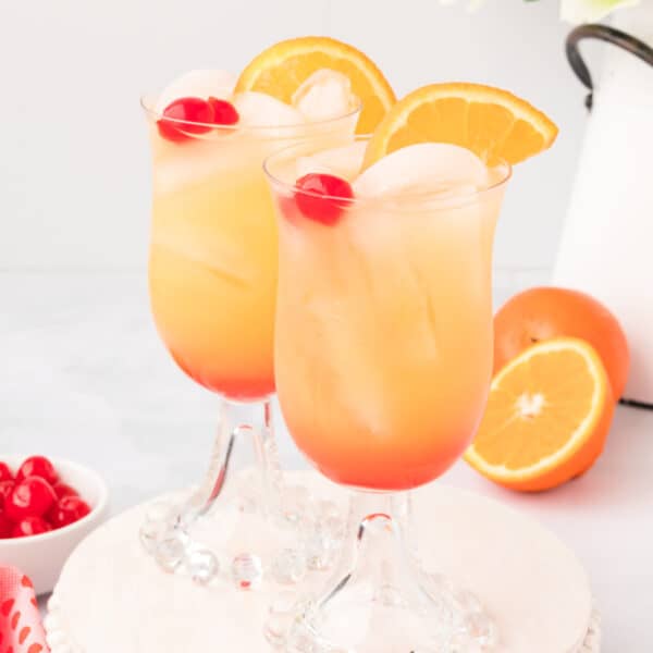 two Arizona Sunset drinks with ice, orange slices, and cherries on a white stand