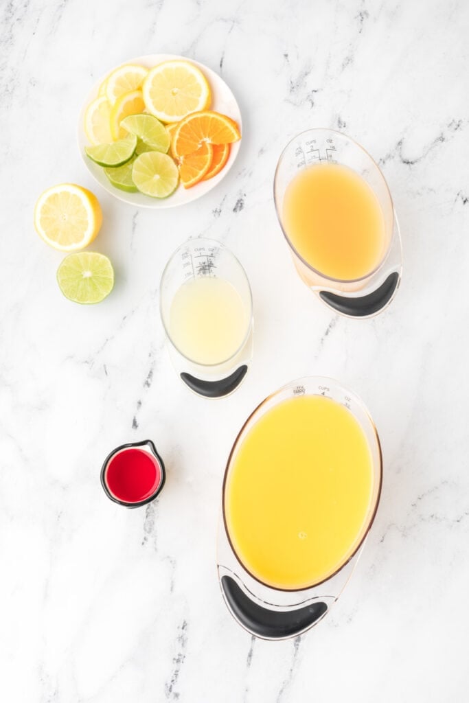 Ingredients for Cactus Cooler recipe including orange juice, pineapple juice, lemon juice, grenadine, and sliced citrus on a white marble surface