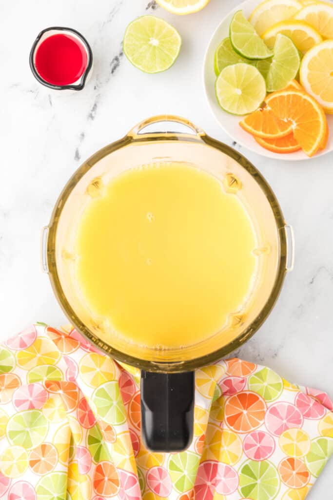 Blender pitcher filled with orange and pineapple juice mixture for homemade Cactus Cooler recipe beside sliced citrus and grenadine