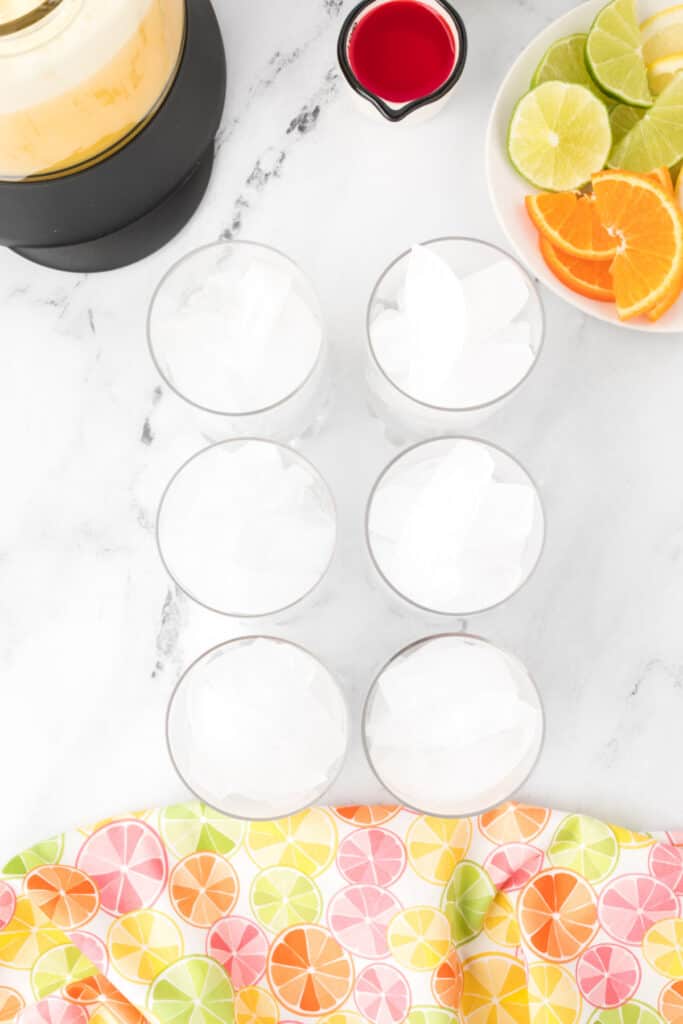 Six glasses filled with ice beside a blender pitcher, sliced citrus, and grenadine for making Cactus Cooler drinks