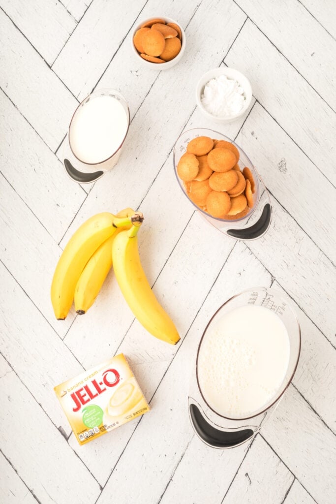 ingredients for banana pudding parfait including bananas, milk, banana cream pudding mix, Nilla wafers, and powdered sugar on a white surface